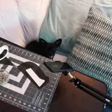 A small black dog watches as someone uses adaptive sewing tools, including a rotary cutter and pins, on a cutting mat.
