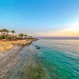 Piękna pogoda w Marsa Alam: piaszczysta plaża z palmami, leżakami i parasolami, turkusowe morze i długie molo na horyzoncie.
