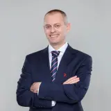 Marcin Włodarski, trener, w granatowym garniturze i krawacie, z rękami skrzyżowanymi na piersi, uśmiecha się do obiektywu.