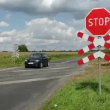 Czarny samochód zbliża się do przejazdu kolejowego z czerwonym znakiem STOP i białoczerwonym krzyżem.