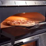 Une pizza garnie est enfournée dans un four à pizza professionnel. La pelle à pizza glisse sous la pâte dorée, prête à être cuite.