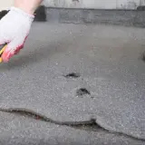 Ręka w rękawicy z nożem przygotowuje dach z papy do naprawy. Widać pęknięcia i dziury, które wymagają interwencji.