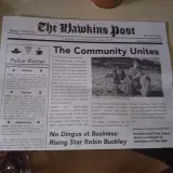 Gazeta "The Hawkins Post" z artykułem "The Community Unites" i zdjęciem dzieci sprzątających plażę.