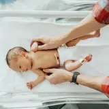 Noworodek w inkubatorze, delikatnie dotykany przez ojca. Widoczny naderwany kikut pępowiny.