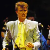 David Bowie na scenie podczas Live Aid, w jasnej marynarce i żółtym krawacie, śpiewa z pasją.