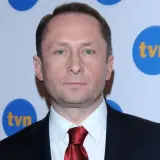 Kamil Durczok w garniturze i czerwonym krawacie na tle logo TVN. Prowadzący Fakty TVN.