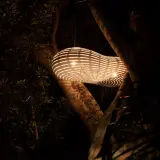 Lámpara colgante de bambú iluminada, suspendida entre árboles oscuros.