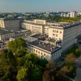 Widok z lotu ptaka na duży kompleks szpitalny z czerwonym krzyżem na fasadzie, otoczony drzewami i parkingiem.