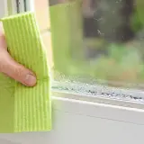 Ręka z zieloną ściereczką wyciera szybę, na której widać krople **kondensacji**.