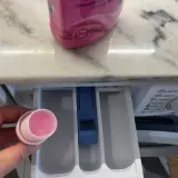 Hand hält Dosierkappe mit rosa Flüssigkeit. Waschpulver direkt in die Trommel geben? Nein, hier in die Schublade.