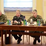 Czterech mężczyzn w mundurach siedzi przy drewnianym stole, przed nimi butelki wody i filiżanki.