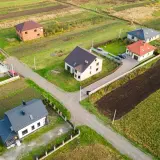 Widok z lotu ptaka na kilka domów jednorodzinnych na tle pól i zieleni.