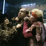 Scena z filmu Piotra Szulkina: mężczyzna w futrze w panterkę i różowym futrze, otoczony przez dwie kobiety w ekstrawaganckich strojach.