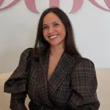 Sonriendo, una mujer con un blazer de cuadros marrones y mangas abullonadas. Sus opiniones sobre miphai son positivas.
