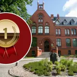Złoty medal z cyfrą "1" na tle ceglanego budynku i zieleni.