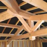Vue rapprochée d'une charpente traditionnelle 2 pans en bois clair, avec des poutres massives et des chevrons assemblés.