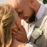 Agnieszka Kaczorowska i jej mąż w czułym uścisku podczas ślubu. Ona ma blond włosy splecione w warkocz, on w jasnej marynarce z butonierką.