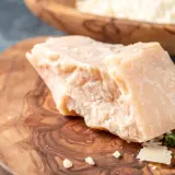 Kawałek twardego sera, przypominającego Grana Padano i parmezan, leży na drewnianej desce. W tle widać misę z tartym serem.