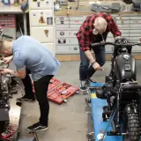 Due uomini lavorano su due moto in un garage. Uno è chino su una moto bianca, l'altro su una nera. Un kit di attrezzi è aperto sul pavimento. Progetto schienalino moto fai da te.
