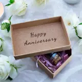 Otwarta szkatułka z napisem "Happy Anniversary" i małą buteleczką w środku, otoczona białymi różami i płonącą świecą. Idealny prezent na 21 rocznicę ślubu.