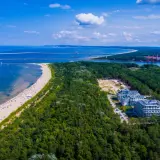 Plaża w Świnoujściu z lotu ptaka. Tłumy plażowiczów, turkusowe morze i zielony las otaczający nowoczesne apartamenty.