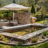 Ein schöner Garten mit einem teil-eingegrabenen Holzpool, Liegestühlen und einer gemütlichen Lounge-Ecke unter einem Sonnenschirm.