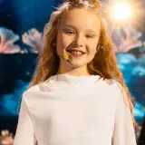 Uśmiechnięta dziewczynka z błyszczącym makijażem na oczach, w białej sukience, gotowa do występu na Eurowizji Junior Polska.