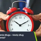 Dłoń trzyma czerwony budzik. Tekst: "Przedawnienie długu – kiedy dług się przedawnia?". Dotyczy to również czynszu najmu.