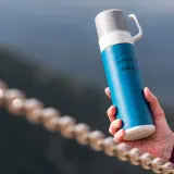 Niebieski termos w góry, z napisem "Mountains are calling" i logo Hi-Tec, trzymany w dłoni na tle górskiego krajobrazu.