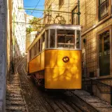 Żółty tramwaj wspina się po stromych torach w wąskiej uliczce Lizbony. W tle widać kamienne budynki pokryte tradycyjnymi płytkami azulejo.