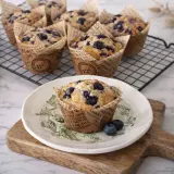 Un délicieux muffin myrtille, tout juste sorti du four, posé sur une assiette décorée. D'autres muffins myrtilles refroidissent sur une grille.