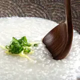 A comforting bowl of Japanese rice porridge (okayu) garnished with fresh scallions and ginger, ready to be enjoyed with a wooden spoon.