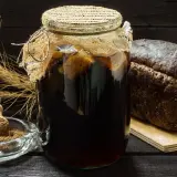 Słoik z domowym kwasem chlebowym, bochenek chleba żytniego, miseczka z mąką i kłosy zboża.