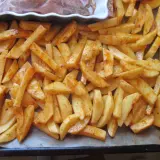 Gotowe do pieczenia frytki w piekarniku, pokrojone ziemniaki doprawione przyprawami, leżą na blasze wyłożonej papierem.