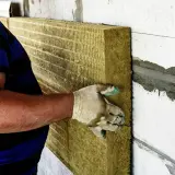 Budowlaniec w rękawicy przykłada płytę z wełny skalnej 10 do ściany.