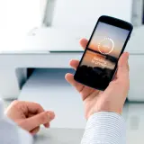 Dłoń trzyma smartfon z napisem "Sending to printer", pokazując jak drukować z telefonu. W tle biała drukarka.