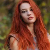 Eine Frau mit langen, dunkelroten Haaren blickt nachdenklich in die Kamera. Ihr Haar fällt ihr über die Schultern, während sie im Freien steht.