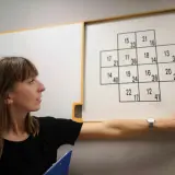 Kobieta wskazuje na tablicę z zadaniami, które mogą być częścią psychotestów. Pytania i odpowiedzi w formie liczb.