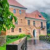 Deszczowy dzień nad zamkiem w Oporowie. Drewniany mostek prowadzi do ceglanego budynku, otoczonego zielenią.