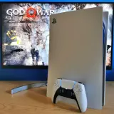 Konsola PlayStation 5 z kontrolerem DualSense obok. Na ekranie widoczny tytuł gry "God of War" i opcje "Nowa gra", "Ustawienia". Idealny moment na playstation aktualizacja.