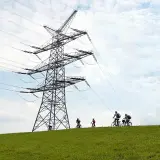 Ludzie na rowerach jadą po zielonym wzgórzu pod ogromną konstrukcją linii wysokiego napięcia.