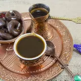 Ein traditionelles Mokkakännchen und eine Tasse mit starkem Mokka-Kaffee, dazu Schokoladenkekse und blaue Blumen auf einem Kupfertablett.