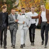 Pięciu mężczyzn w eleganckich strojach na wesele jako goście. Jeden w jasnej, dwurzędowej marynarce, inny w brązowej.