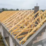 Drewniana konstrukcja dachu bez podbitki, widoczna na budowie domu. Widać komin i elementy ścian z bloczków.