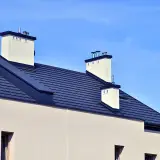 Nowoczesny dach bezokapowy z ciemnogranatową dachówką i białymi kominami na tle błękitnego nieba.