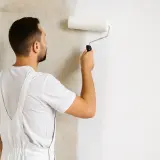 Mann trägt mit einer Rolle Rollputz auf eine Wand auf. Die Wand wird von rauem Beton zu glattem Weiß verändert.