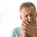Hombre mayor con los ojos cerrados y la mano en la mejilla, sufriendo de dolor, posiblemente por alveolitis seca.