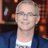 Robert Janowski ile ma lat? Uśmiechnięty mężczyzna w okularach, w granatowej marynarce i białej koszulce.