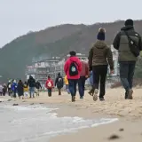 Spacer po plaży, gdy jest długi weekend. Ludzie w ciepłych ubraniach idą brzegiem morza, w tle widać zabudowania i wzgórza.