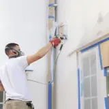 Uomo con maschera protettiva e guanti rossi esegue pittura a spruzzo su una facciata esterna, proteggendo finestre e tubi con nastro adesivo.